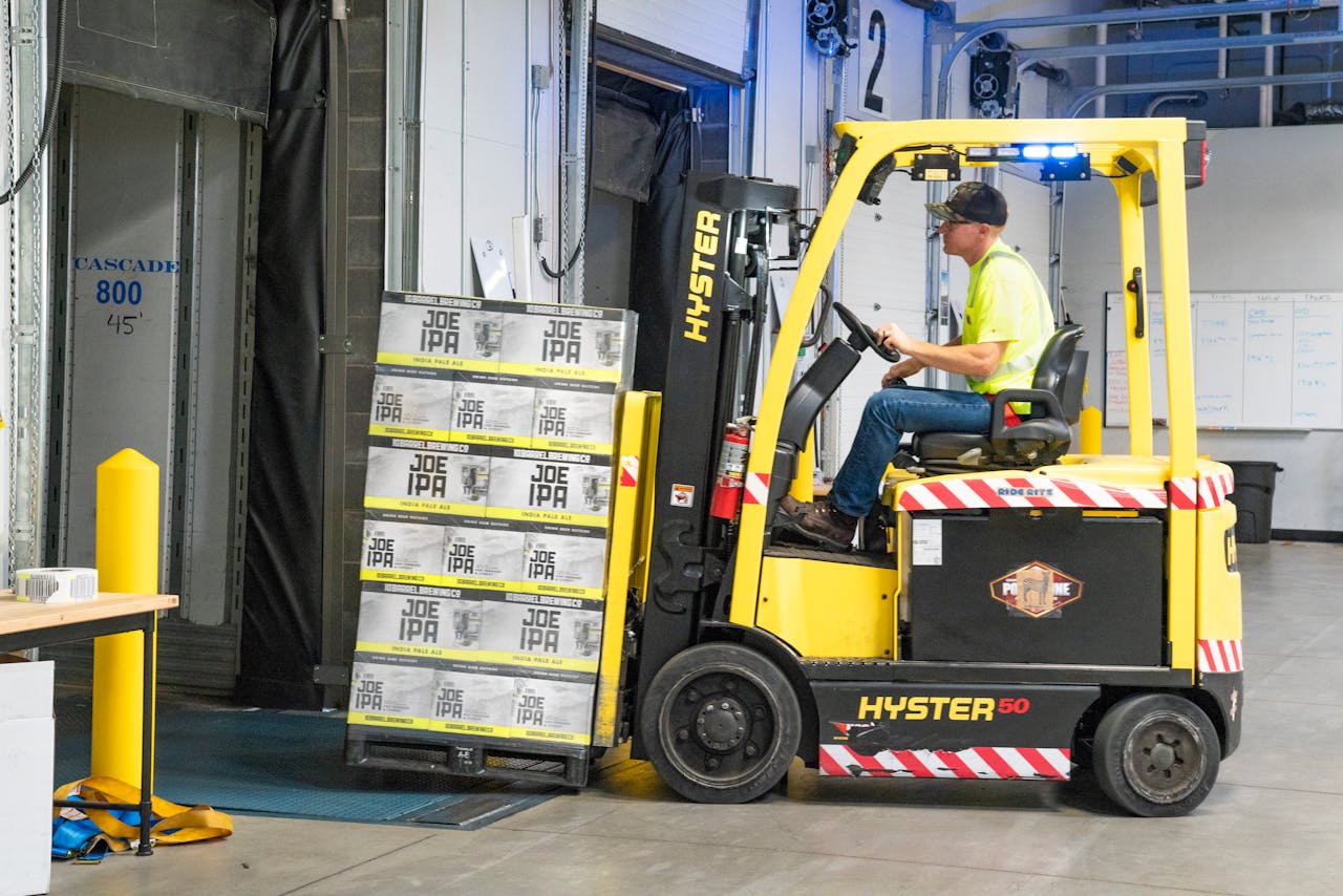 Home A worker drives a Hyster forklift moving Joe IPA boxes in a warehouse.