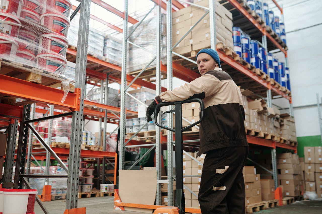 Home Warehouse worker operating a pallet jack among stacked boxes and metal shelves in an industrial setting.