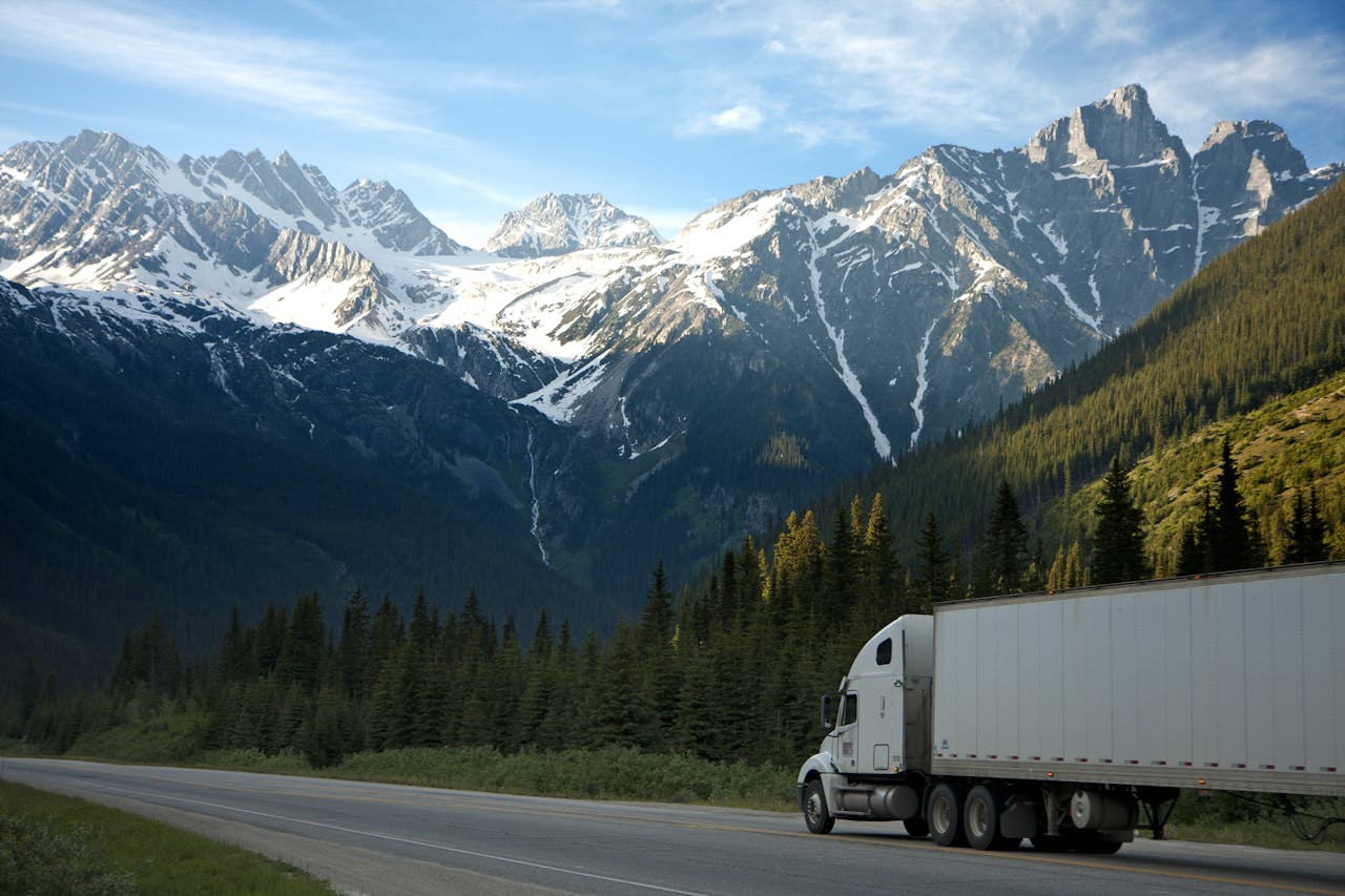 Home A semi-truck travels along a highway with snow-capped mountains in the background.